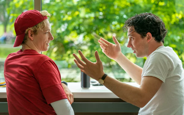 HD wallpaper from the movie Challengers (2024) showing two men engaged in an animated conversation beside a window.