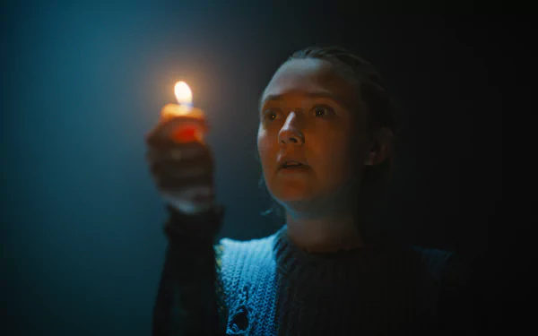 HD wallpaper of Dakota Fanning in The Watchers (2024), holding a lit candle in a dark setting, illuminating her face with a soft glow.