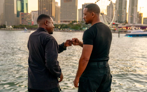 HD wallpaper from Bad Boys: Ride or Die featuring Will Smith and Martin Lawrence fist-bumping at a waterfront with a cityscape in the background.