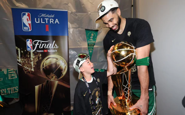 NBA player Jayson Tatum of the Boston Celtics joyfully interacts with a young fan while holding the championship trophy. The image captures a memorable moment from the NBA Finals.