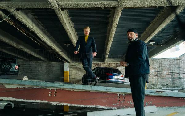 HD desktop wallpaper featuring a scene from the movie Kinds of Kindness. Two people stand in an urban, industrial setting under an overpass, with one leaning on a railing and the other standing nearby.