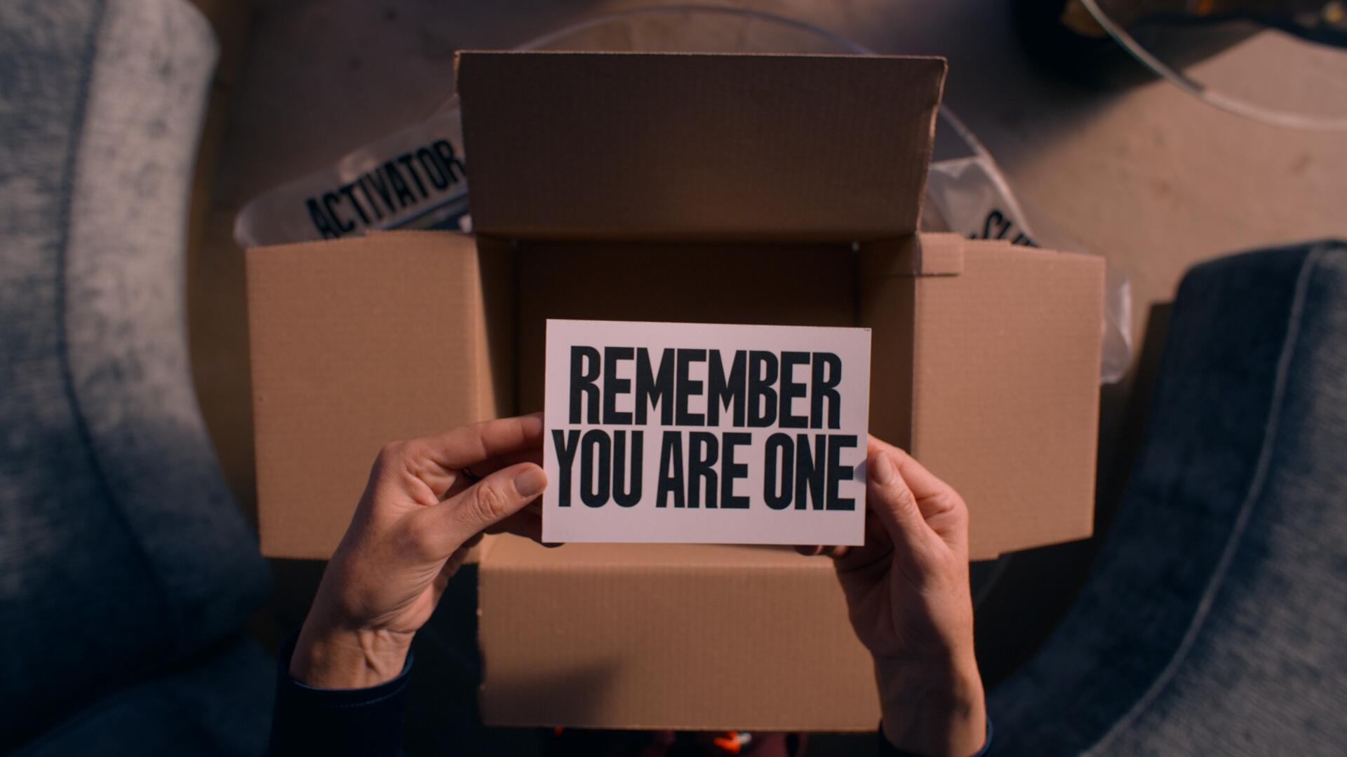 A still from the movie The Substance, featuring hands holding a card that reads REMEMBER YOU ARE ONE, set against a backdrop of an open cardboard box.
