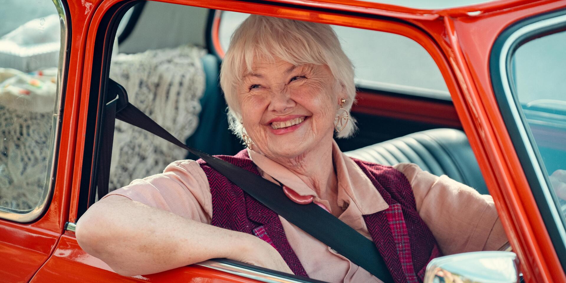 Smiling elderly woman seated in a vintage red car, featured in a 4K Ultra HD Heartstopper TV show desktop wallpaper.