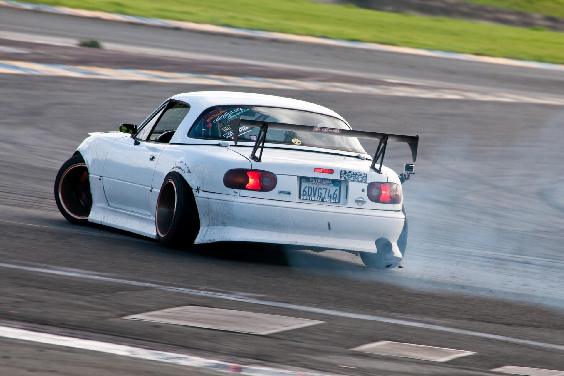 A Mazda MX-5 drifts on a racetrack, showcasing its JDM style with a sleek white body and large rear wing, surrounded by smoke and excitement. HD wallpaper for car enthusiasts.