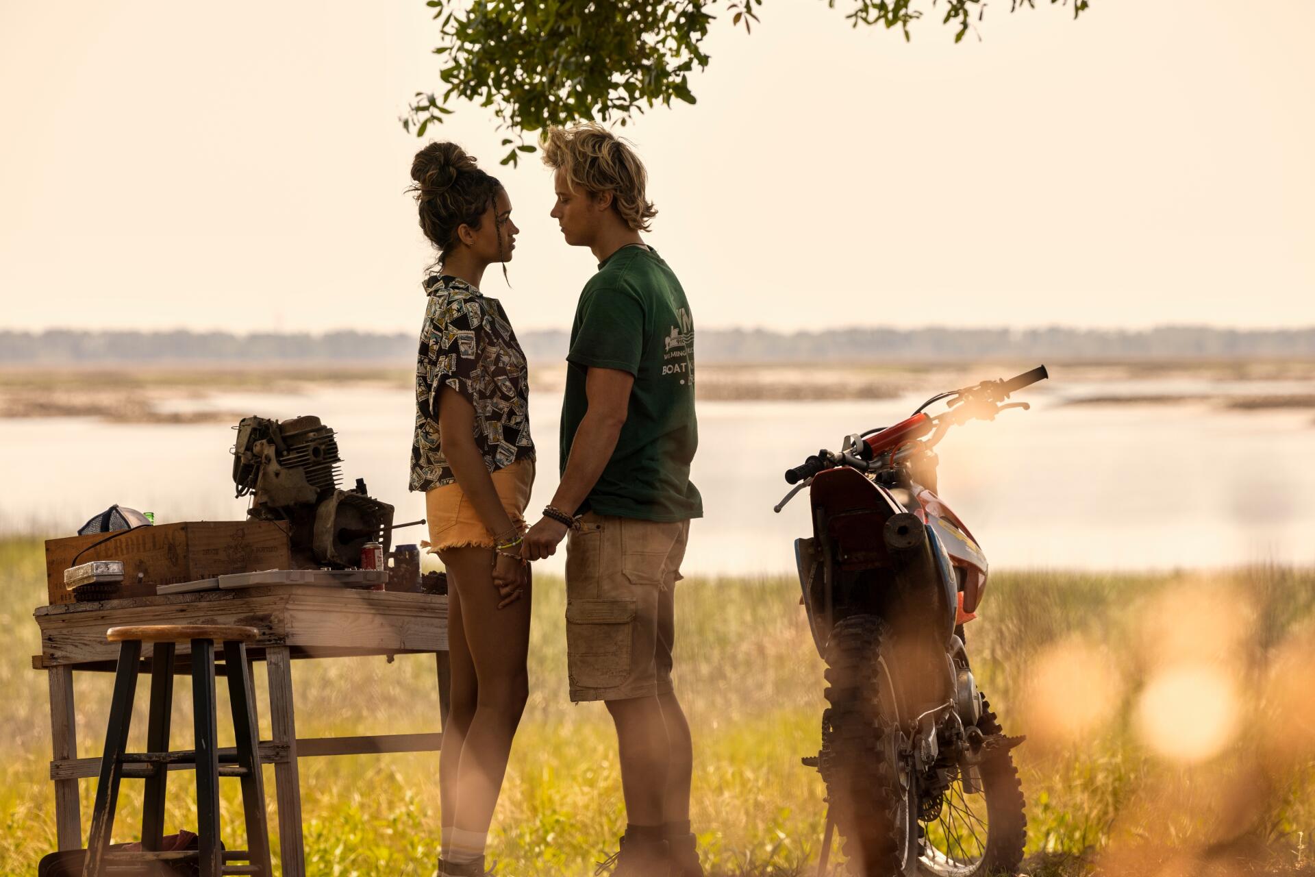 A serene outdoor scene from the TV show Outer Banks, featuring two characters sharing a moment while holding hands, with a scenic background of water and greenery.