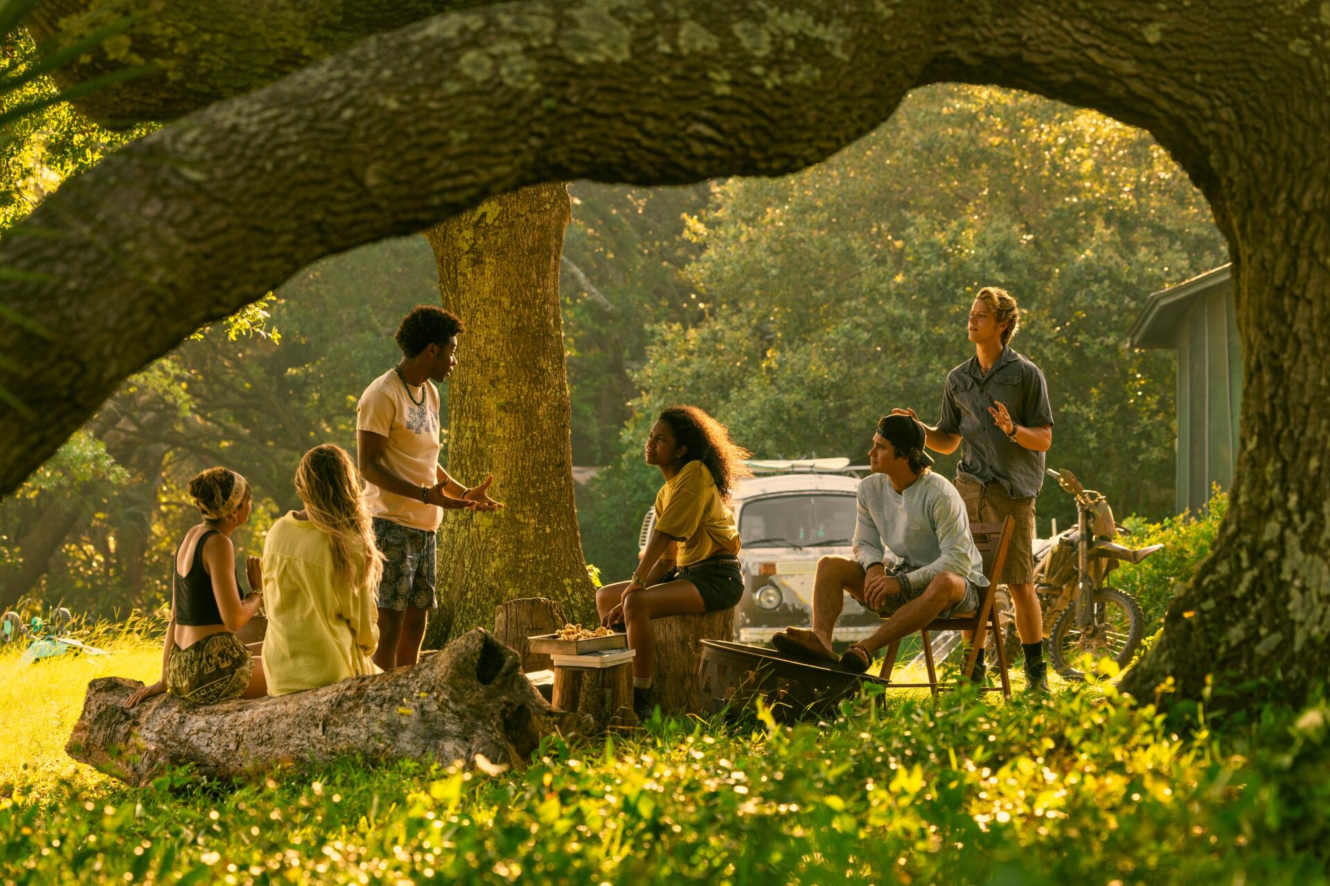 A scenic moment from the TV show Outer Banks, featuring a group of friends gathered under a tree, enjoying each other's company with a vintage car in the background.