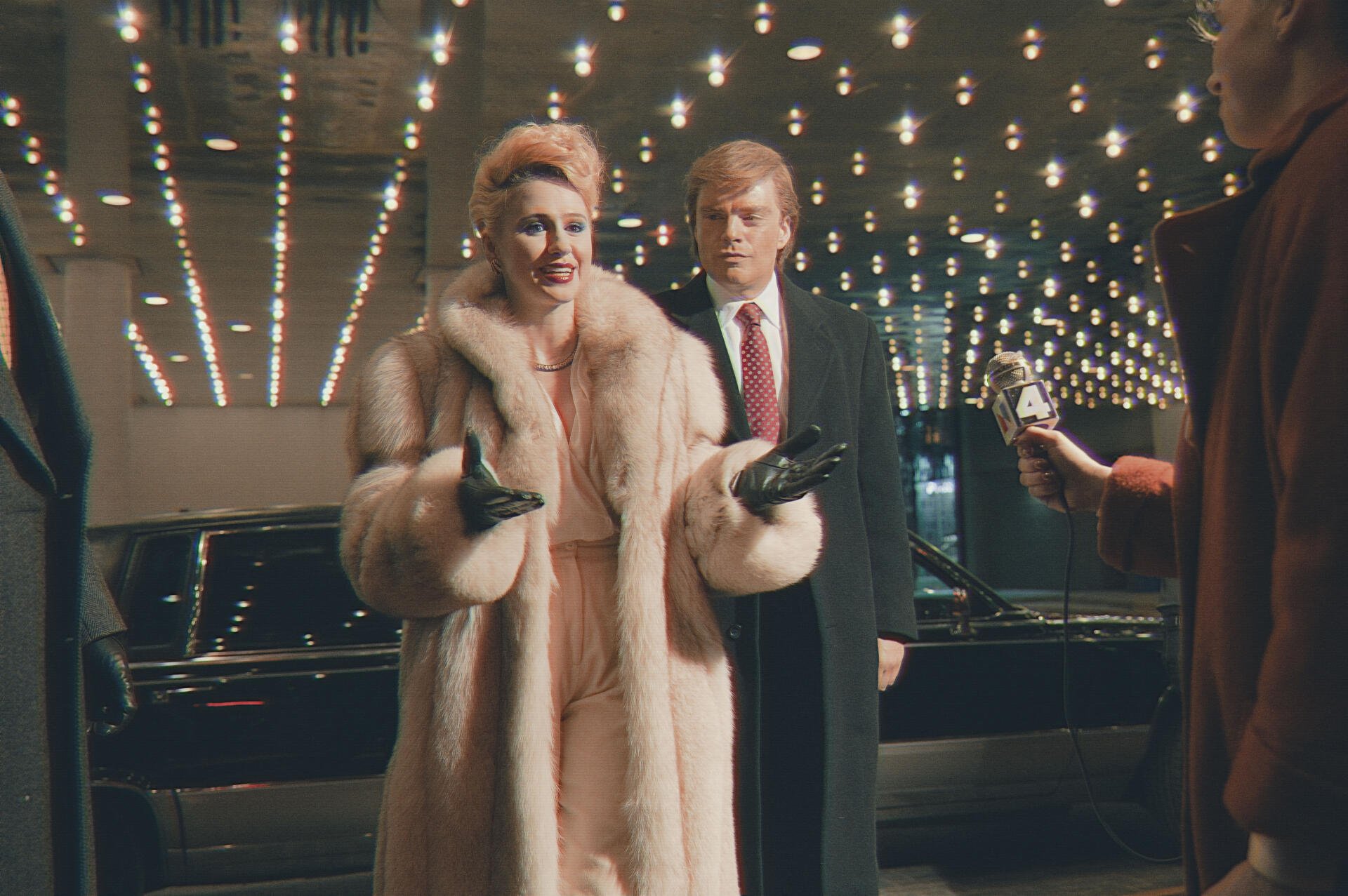 A scene from *The Apprentice (2024)* featuring a glamorous character in a fur coat and a suited figure amidst dazzling lights, set against a sleek black car backdrop. 4K Ultra HD wallpaper.