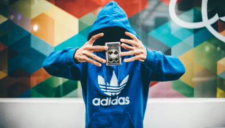 A hooded figure in an Adidas hoodie performs a magic trick with a deck of cards against a vibrant, geometric background. This HD image captures a blend of style and mystique.