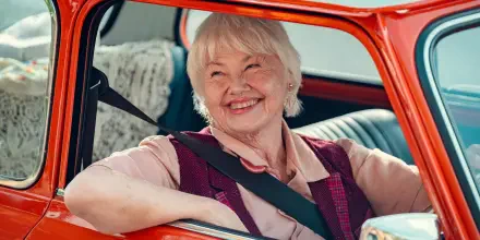 Smiling elderly woman seated in a vintage red car, featured in a 4K Ultra HD Heartstopper TV show desktop wallpaper.