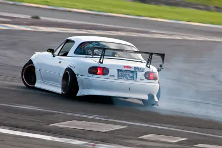 A Mazda MX-5 drifts on a racetrack, showcasing its JDM style with a sleek white body and large rear wing, surrounded by smoke and excitement. HD wallpaper for car enthusiasts.