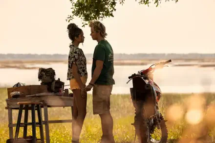 A serene outdoor scene from the TV show Outer Banks, featuring two characters sharing a moment while holding hands, with a scenic background of water and greenery.