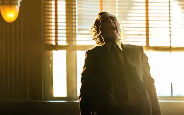 Joaquin Phoenix as the Joker in Joker: Folie à Deux, captured in a dramatic moment with sunlight filtering through blinds, creating a striking 4K Ultra HD wallpaper.