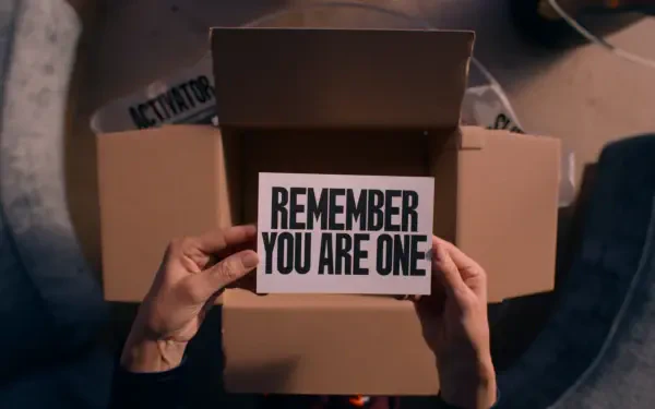 A still from the movie The Substance, featuring hands holding a card that reads REMEMBER YOU ARE ONE, set against a backdrop of an open cardboard box.