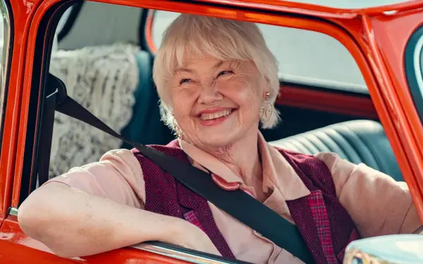 Smiling elderly woman seated in a vintage red car, featured in a 4K Ultra HD Heartstopper TV show desktop wallpaper.