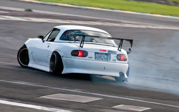 A Mazda MX-5 drifts on a racetrack, showcasing its JDM style with a sleek white body and large rear wing, surrounded by smoke and excitement. HD wallpaper for car enthusiasts.
