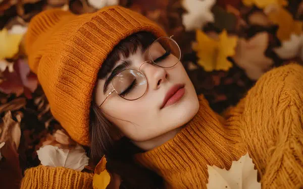 A woman in a cozy orange beanie and sweater lies among autumn leaves, exuding a warm, serene vibe. This 4K Ultra HD image captures the essence of a cozy autumn aesthetic.