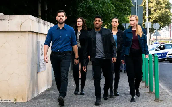 A group of five agents from the TV show FBI: International confidently walks down a city street, showcasing determination and teamwork against a vibrant urban backdrop.