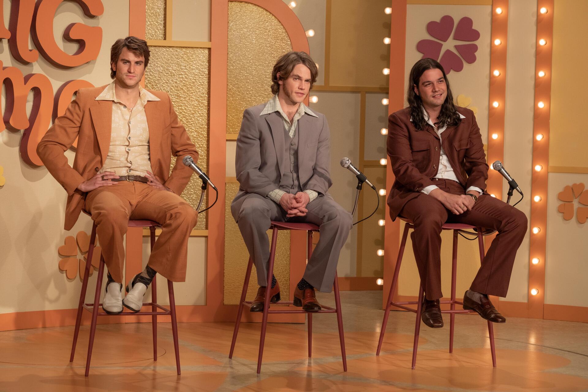 Three men seated on tall stools in a retro game show setting, dressed in stylish outfits, from the movie Woman of the Hour. This vibrant image serves as a 4K Ultra HD desktop wallpaper.