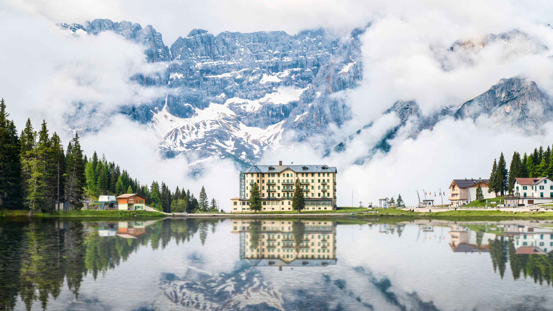 A serene landscape featuring a grand mansion by a lake, its reflection mirrored in the calm water, surrounded by majestic mountains and soft clouds overhead. 4K Ultra HD wallpaper.