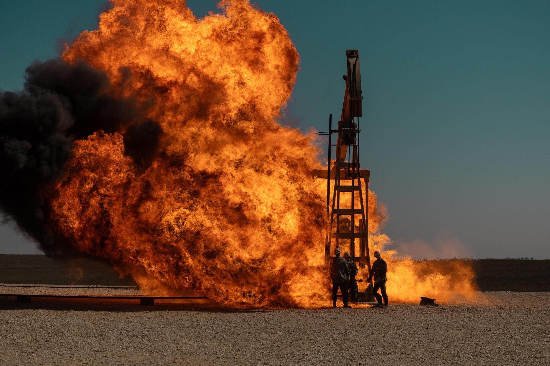 A dramatic scene from the TV show Landman, featuring a towering explosion and flames against a clear blue sky, capturing intense action and tension.