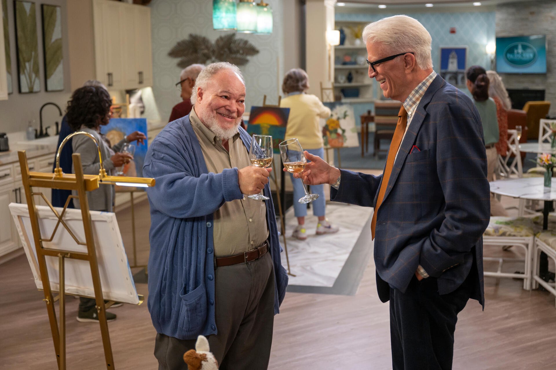 A scene from the TV show A Man on the Inside featuring Ted Danson and Stephen McKinley Henderson sharing a toast, set in a vibrant community space.