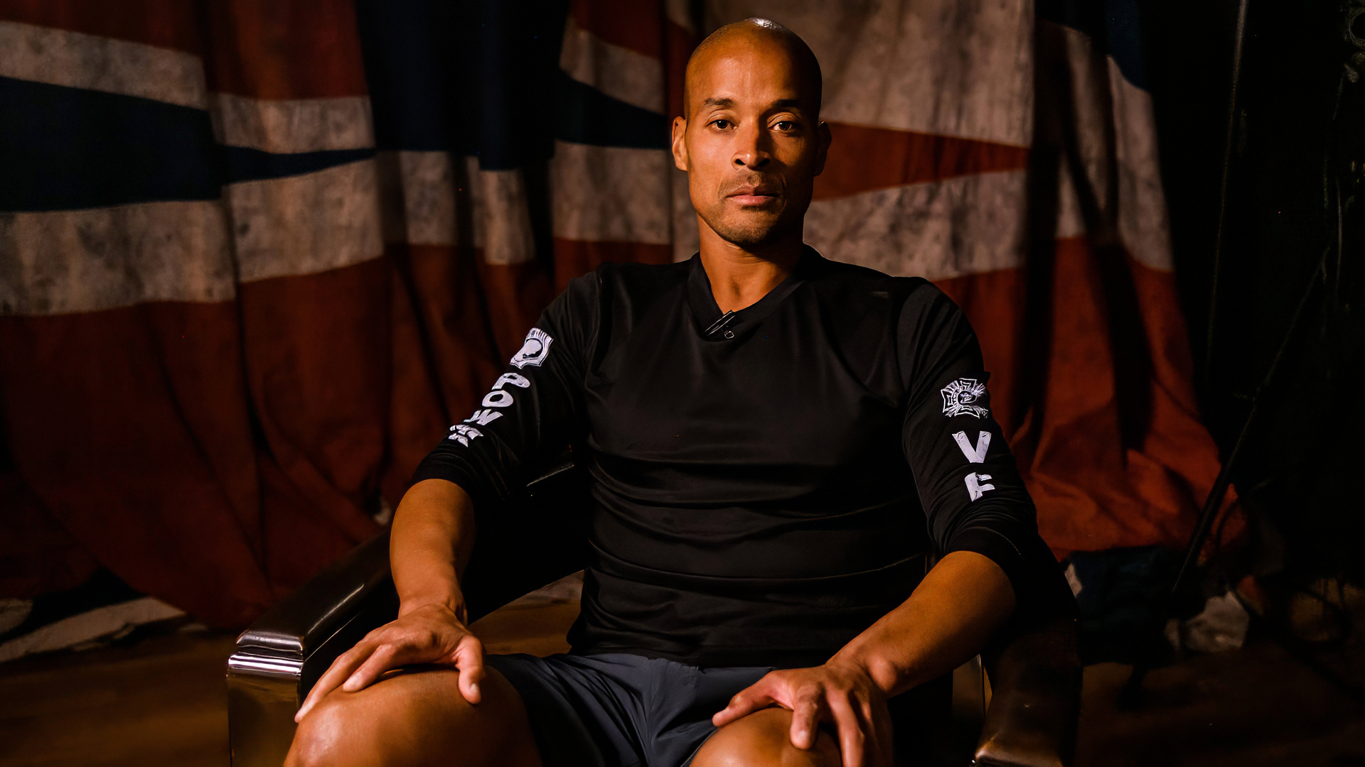 David Goggins sits confidently in front of a flag backdrop, embodying determination and strength. This HD wallpaper captures his inspiring presence in sports.