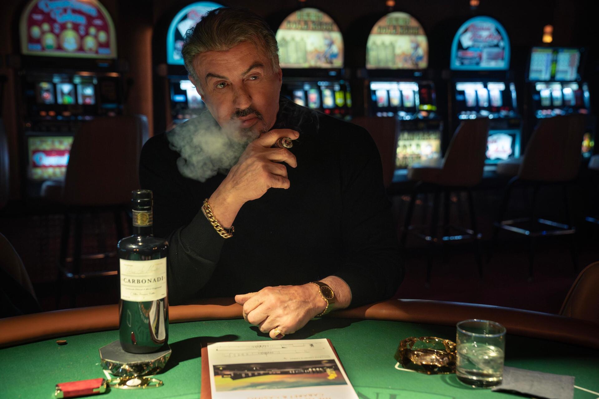 A scene from Tulsa King features Sylvester Stallone, smoking cigar and enjoying whiskey at a casino table, with colorful slot machines in the background. HD wallpaper for PC.