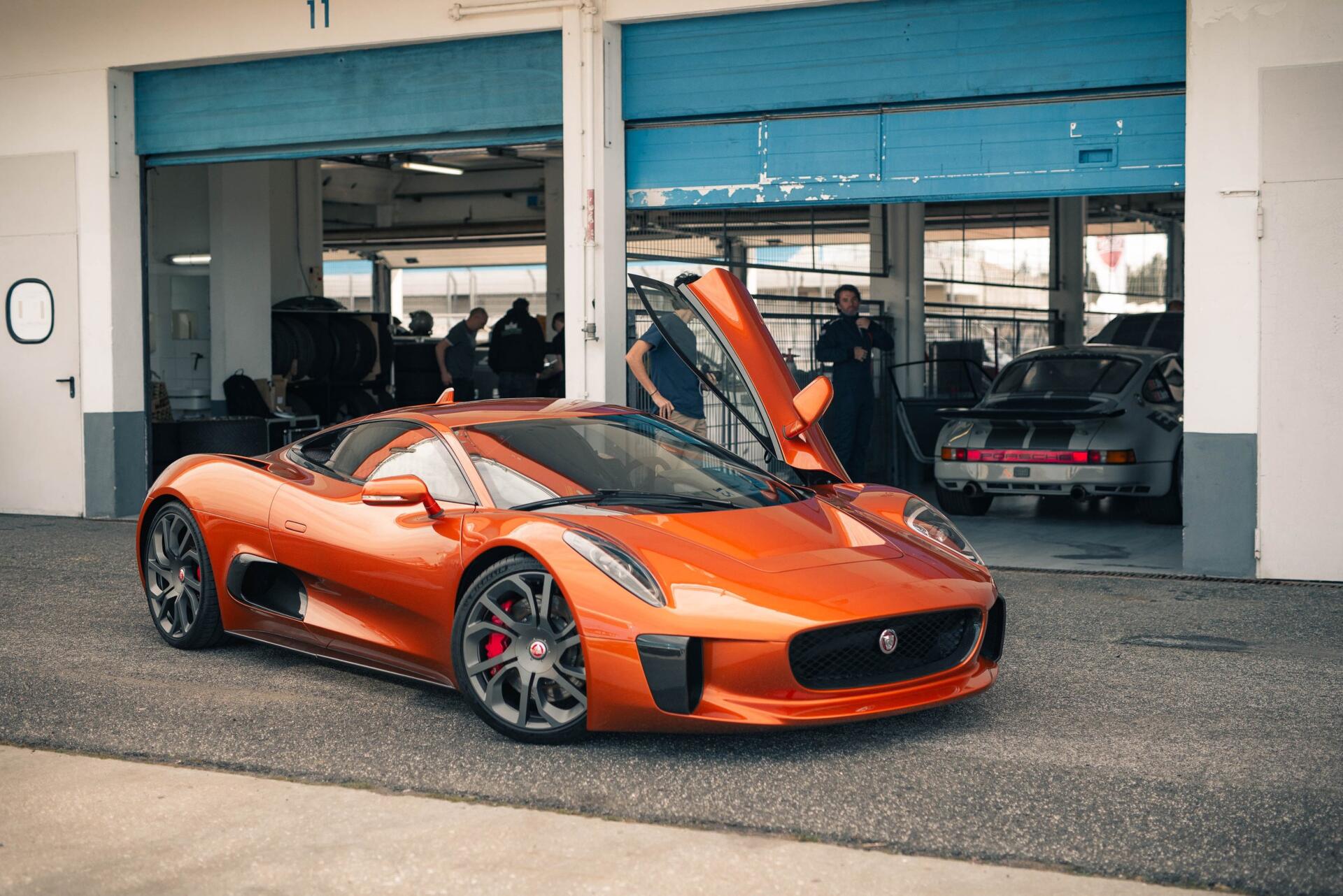 An orange Jaguar C-X75 luxury car with its doors open, parked in front of a garage with other cars visible in the background. A striking HD wallpaper for automotive enthusiasts.