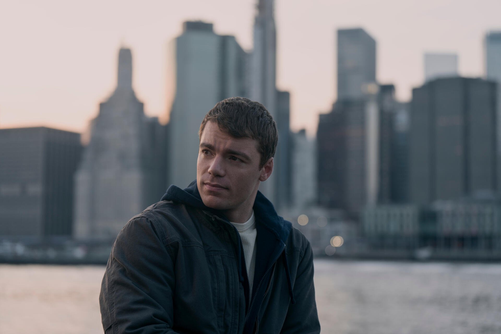 Gabriel Basso in a scene from the TV show The Night Agent, seated against a backdrop of a city skyline during twilight, creating a dramatic and engaging atmosphere.