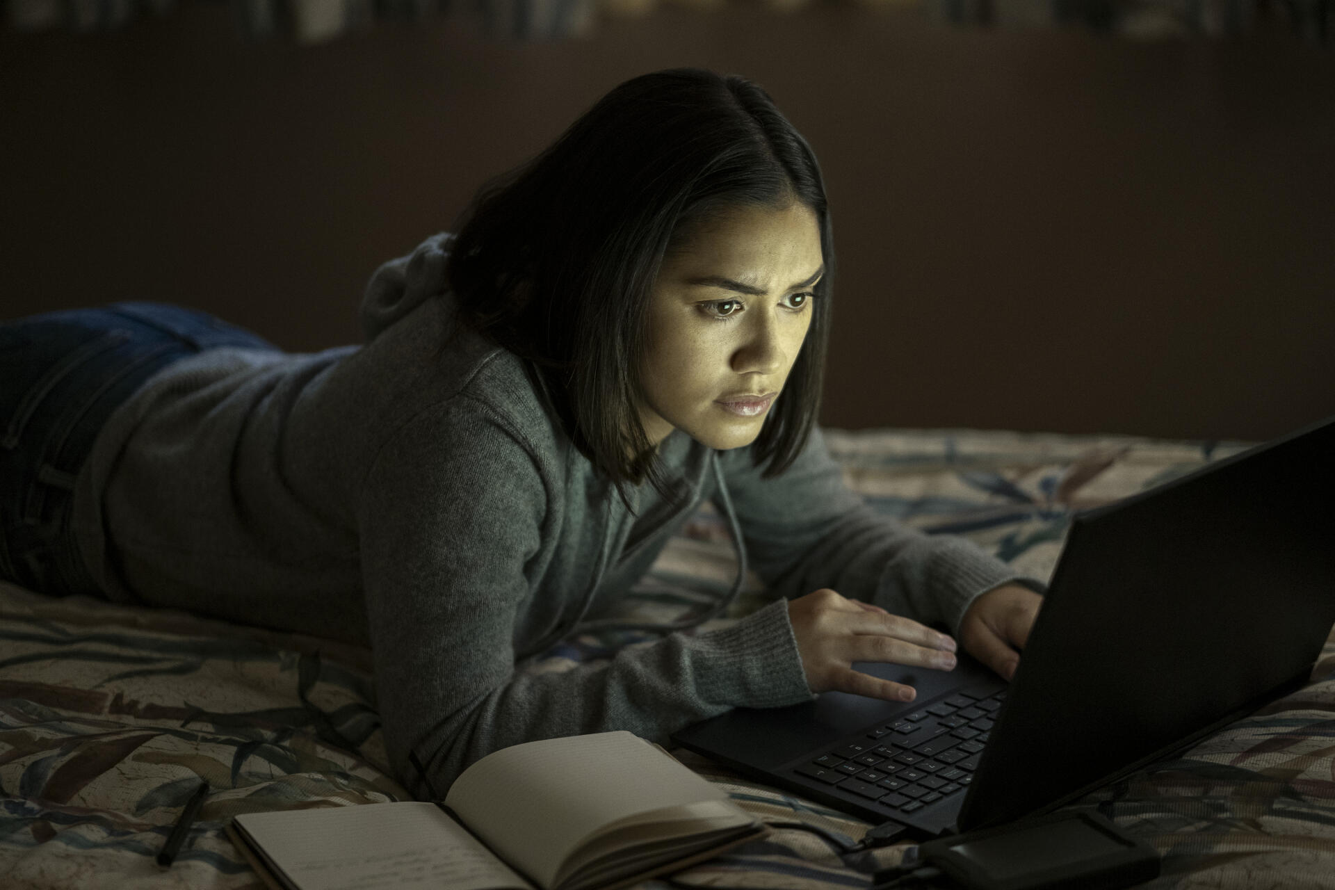 A scene from the TV show The Night Agent, featuring Luciane Buchanan deeply focused on her laptop while resting on a bed, captured in a dark, atmospheric setting.