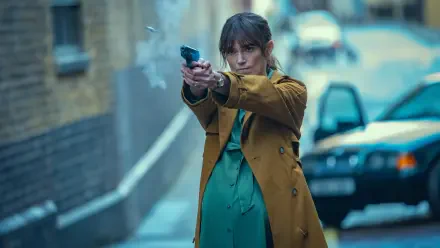 Keira Knightley in a tense scene from the TV show Black Doves, aiming a gun while wearing a brown coat and a green dress, set against an urban backdrop. 4K Ultra HD desktop wallpaper.