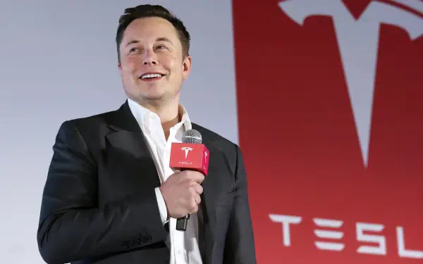 Elon Musk smiles while holding a microphone at a Tesla event, set against a vibrant Tesla logo background, creating an engaging HD desktop wallpaper.