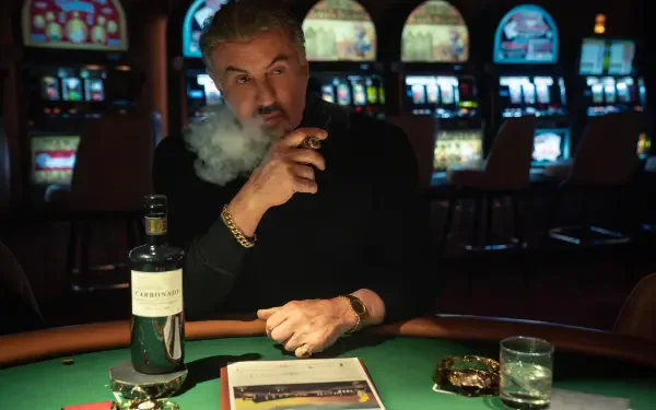A scene from Tulsa King features Sylvester Stallone, smoking cigar and enjoying whiskey at a casino table, with colorful slot machines in the background. HD wallpaper for PC.