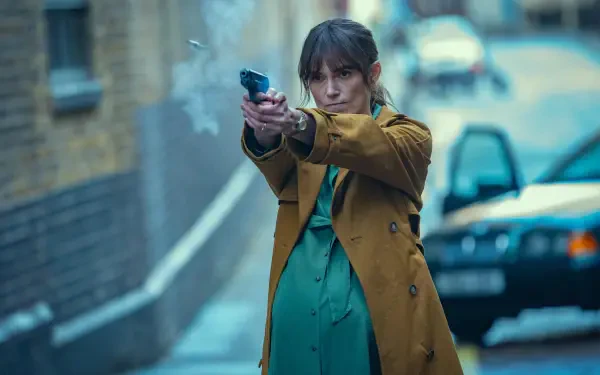 Keira Knightley in a tense scene from the TV show Black Doves, aiming a gun while wearing a brown coat and a green dress, set against an urban backdrop. 4K Ultra HD desktop wallpaper.