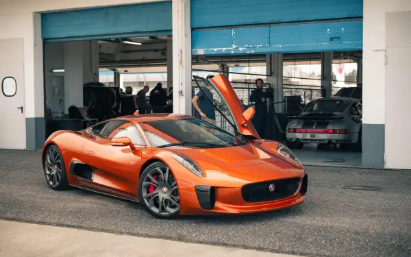 An orange Jaguar C-X75 luxury car with its doors open, parked in front of a garage with other cars visible in the background. A striking HD wallpaper for automotive enthusiasts.