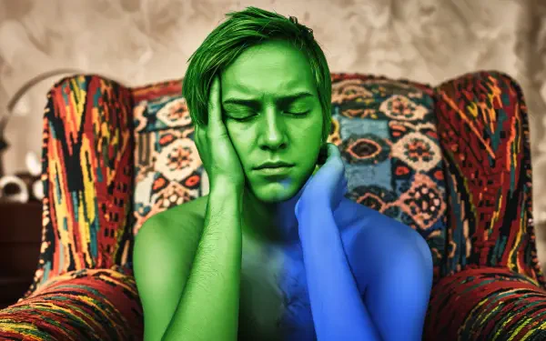 A tired individual rests with their head in their hands, seated in a colorful armchair. Their skin is artistically tinted in green and blue, creating a striking effect in this 4K Ultra HD wallpaper.