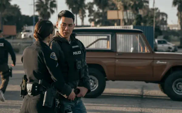 A scene from the TV show On Call (2025) featuring two police officers engaged in conversation, with a vintage brown vehicle and palm trees in the background. 4K Ultra HD wallpaper.