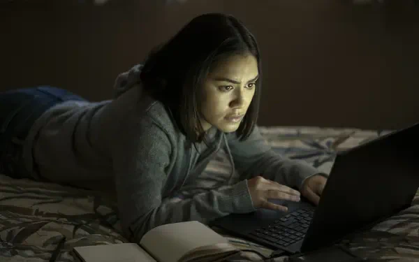 A scene from the TV show The Night Agent, featuring Luciane Buchanan deeply focused on her laptop while resting on a bed, captured in a dark, atmospheric setting.