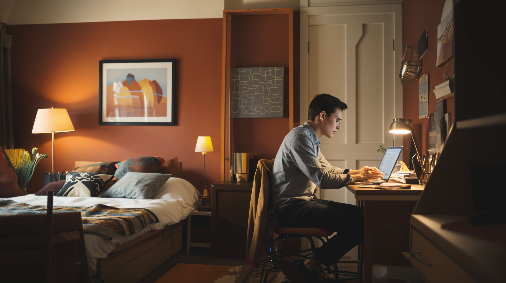 A digital nomad works focused at a desk in a cozy, warmly-lit room, with a bed and artwork in the background, showcasing a blend of productivity and comfort.