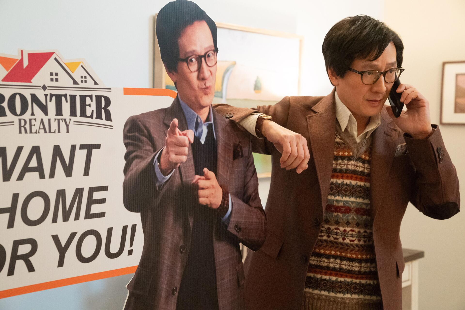 A scene from the movie Love Hurts, featuring a character in a brown suit standing next to a life-sized cardboard cutout, holding a phone and interacting with a Frontier Realty sign.