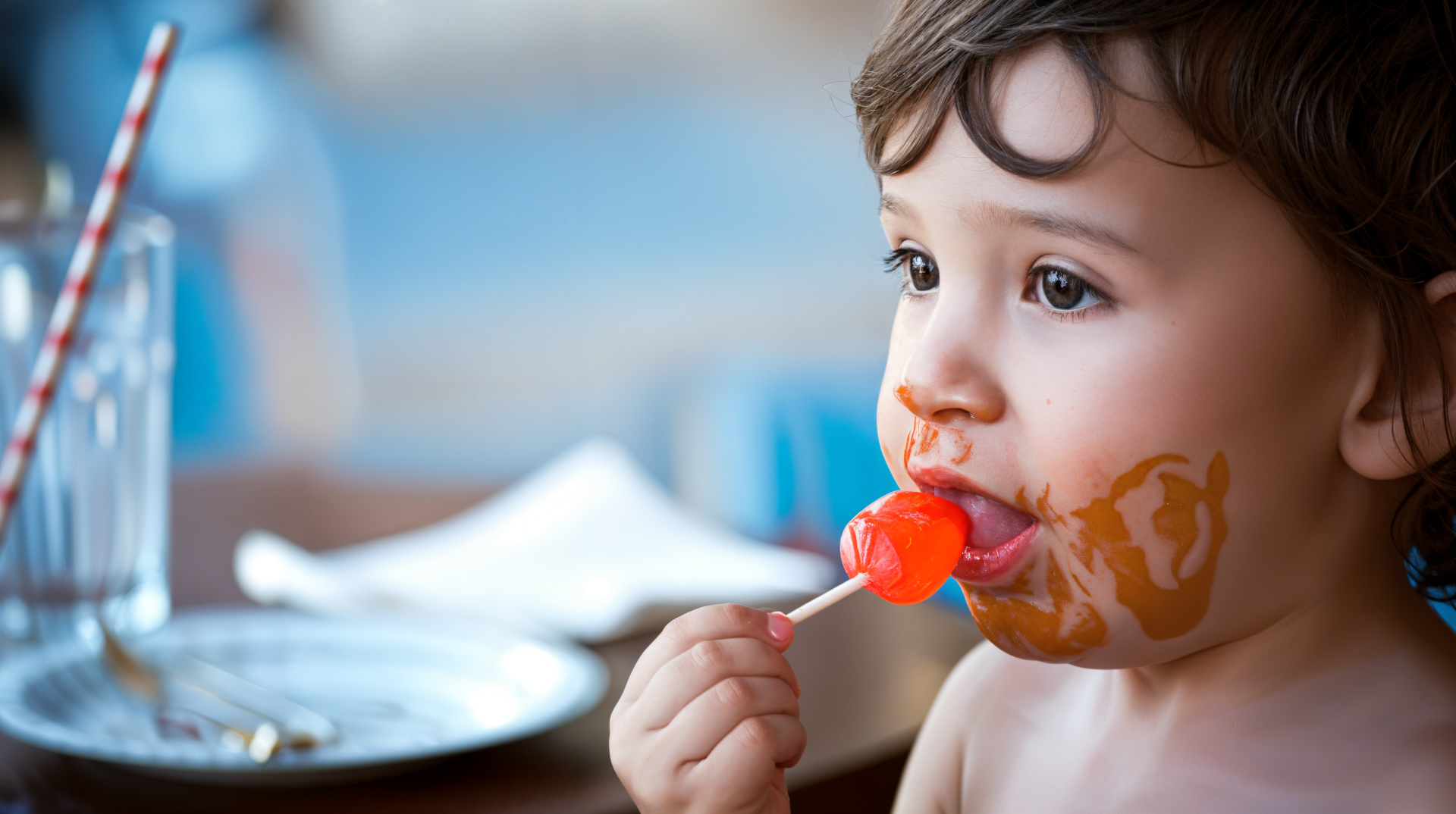 A young child enjoys a bright red lollipop, with playful expression and a smeared candy face, set against a softly blurred background, captured in vibrant 4K Ultra HD.
