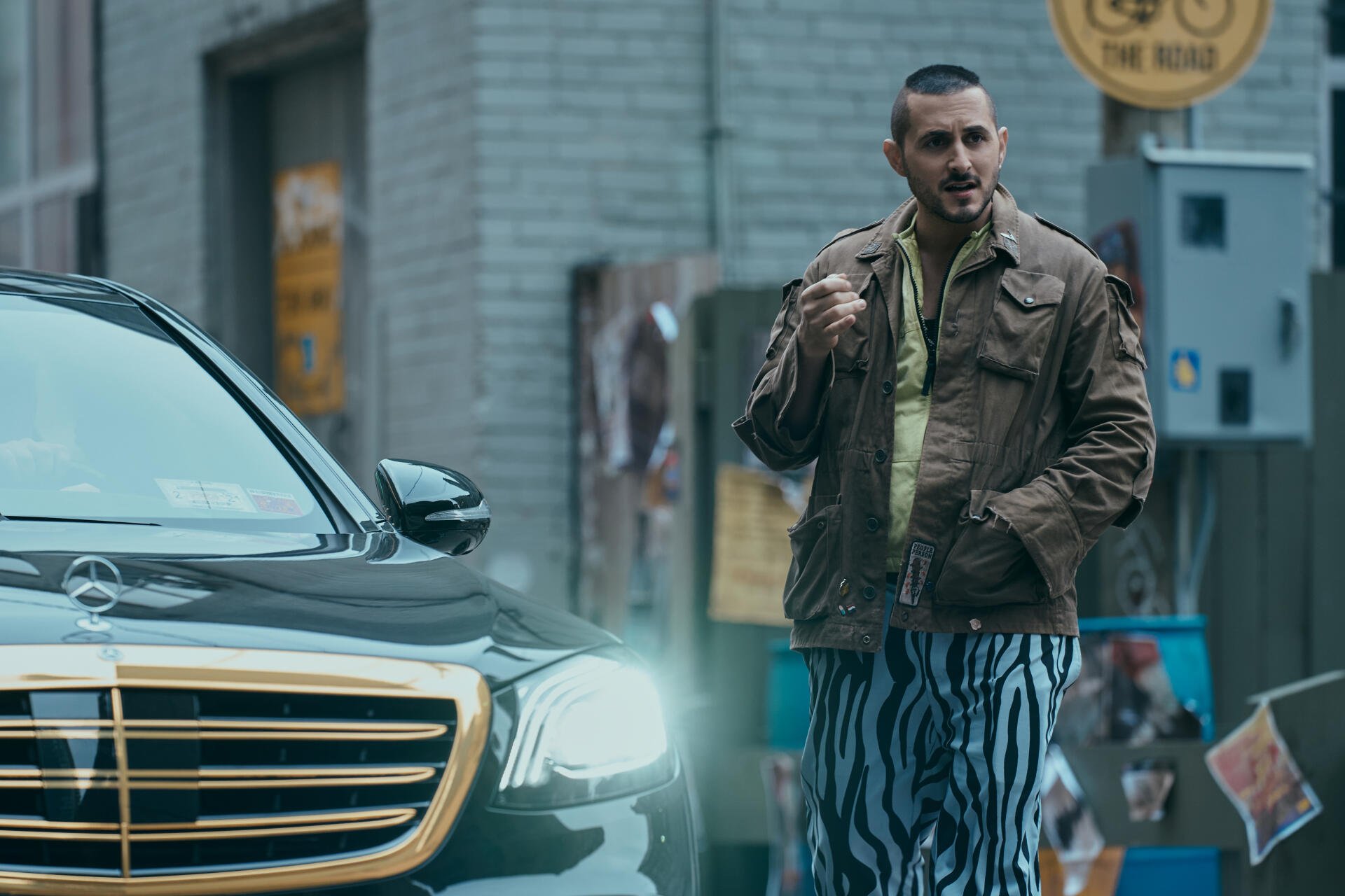 A scene from The Boys (2019) featuring a character in a brown jacket and zebra-striped pants, standing next to a sleek black car in an urban setting. 4K Ultra HD quality.
