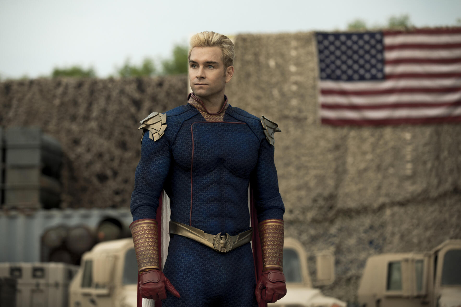 Homelander from the TV show The Boys stands confidently in a military setting, with an American flag in the background, showcasing his powerful presence as a key character from the series.
