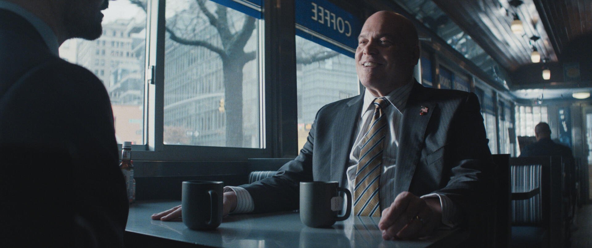 A scene from Daredevil: Born Again, featuring a man in a suit sitting at a café table with two coffee mugs, showcasing a thoughtful conversation in a modern urban setting.