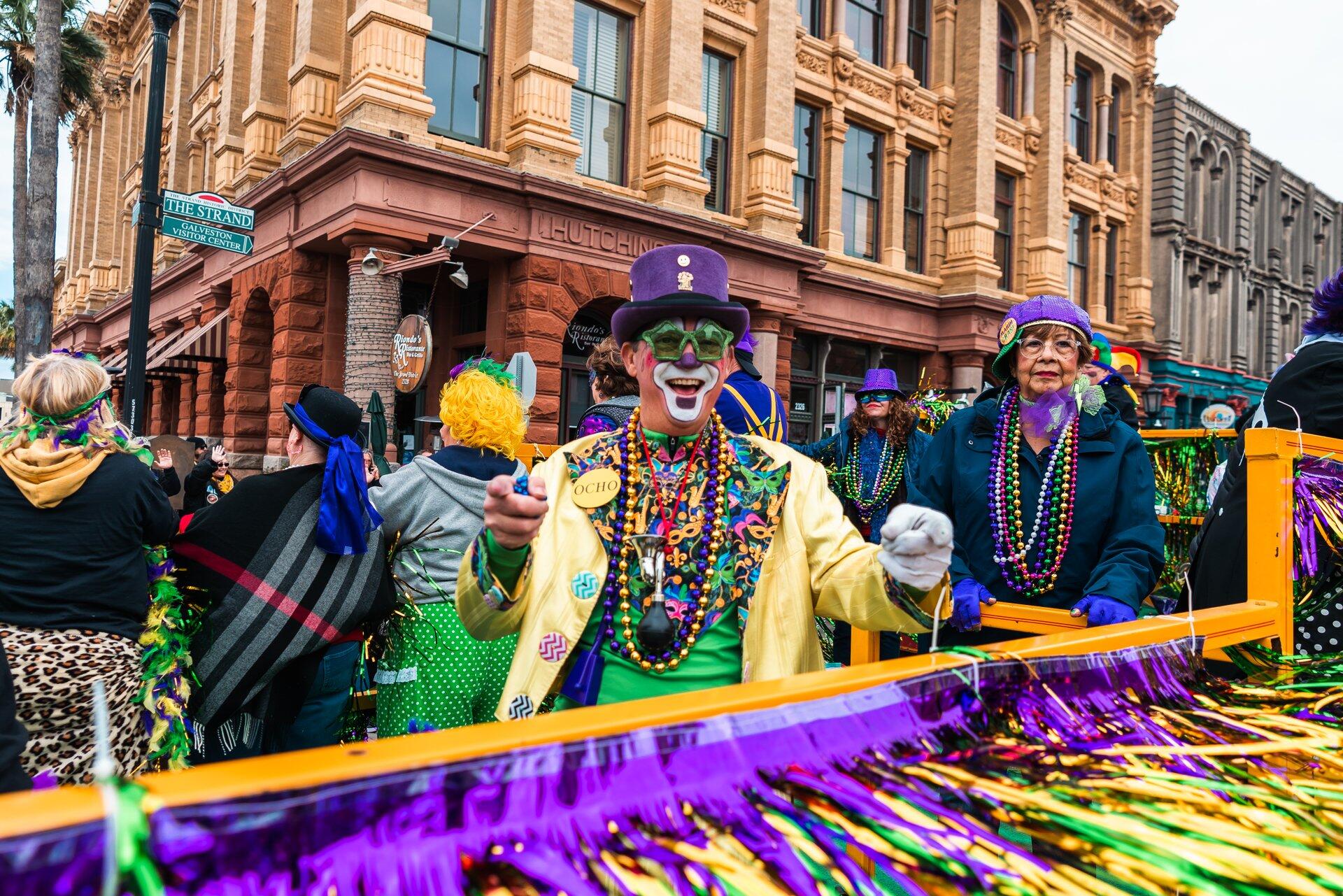 A vibrant Mardi Gras festival scene featuring costumed participants adorned in beads and masks, set against festive decorations and historic architecture. HD wallpaper showcasing celebration.