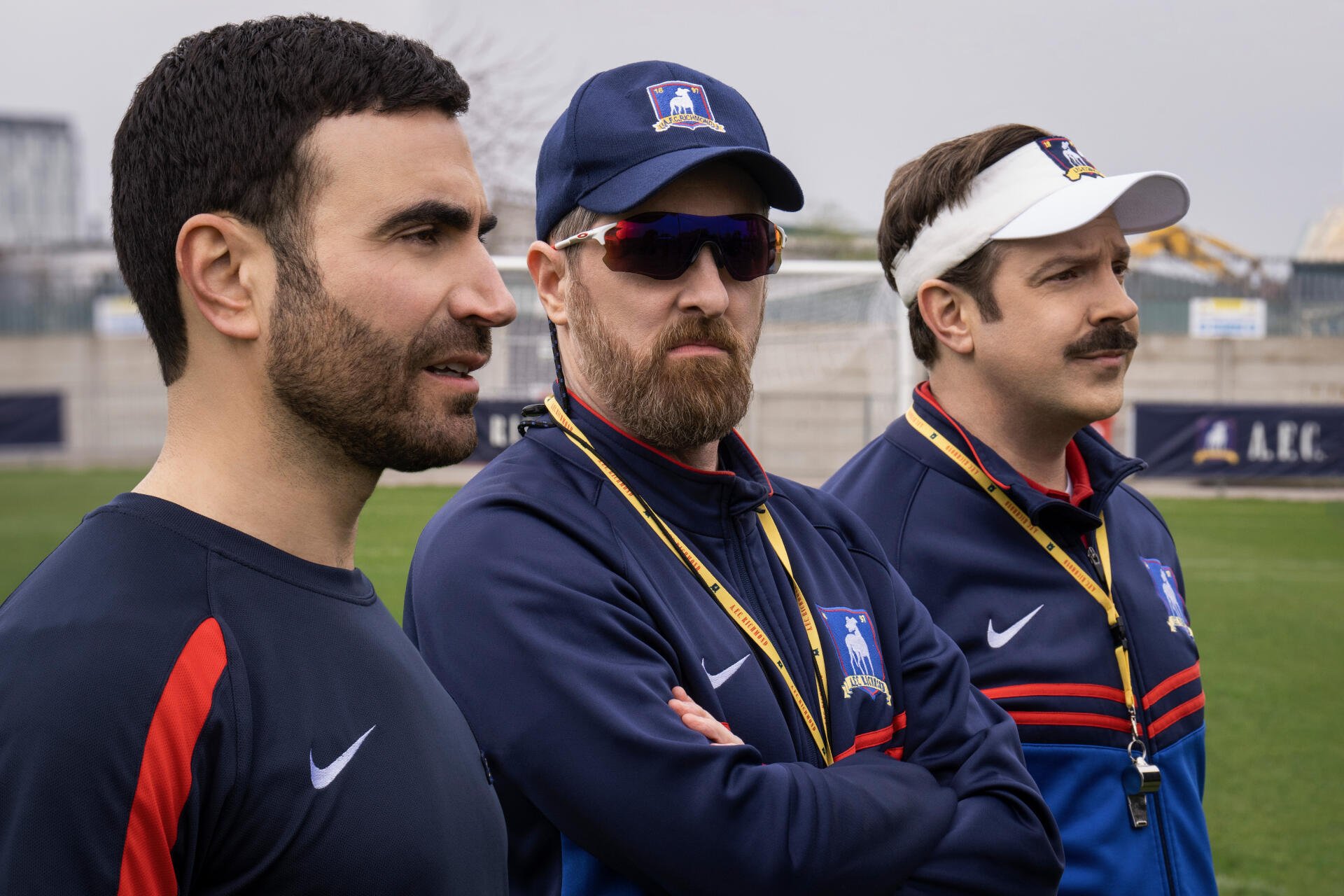 A scene from the TV show Ted Lasso, featuring Jason Sudeikis, Brendan Hunt, and Brett Goldstein as they observe a practice on the field, portrayed in vibrant 8K Ultra HD.