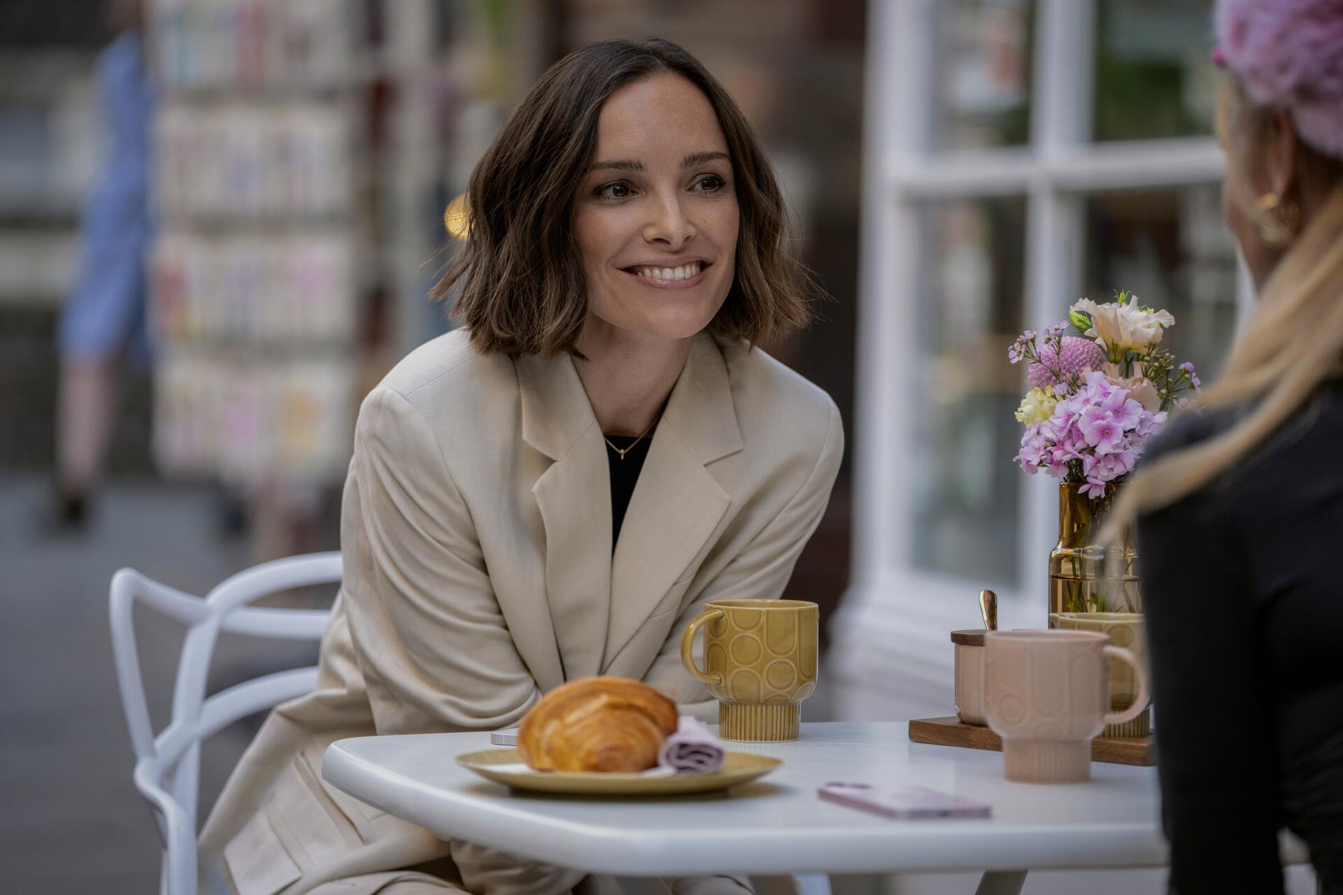 A scene from the TV show Ted Lasso featuring Jodi Balfour, smiling at a table with coffee and pastries, surrounded by flowers in an inviting café setting. 8K Ultra HD wallpaper.