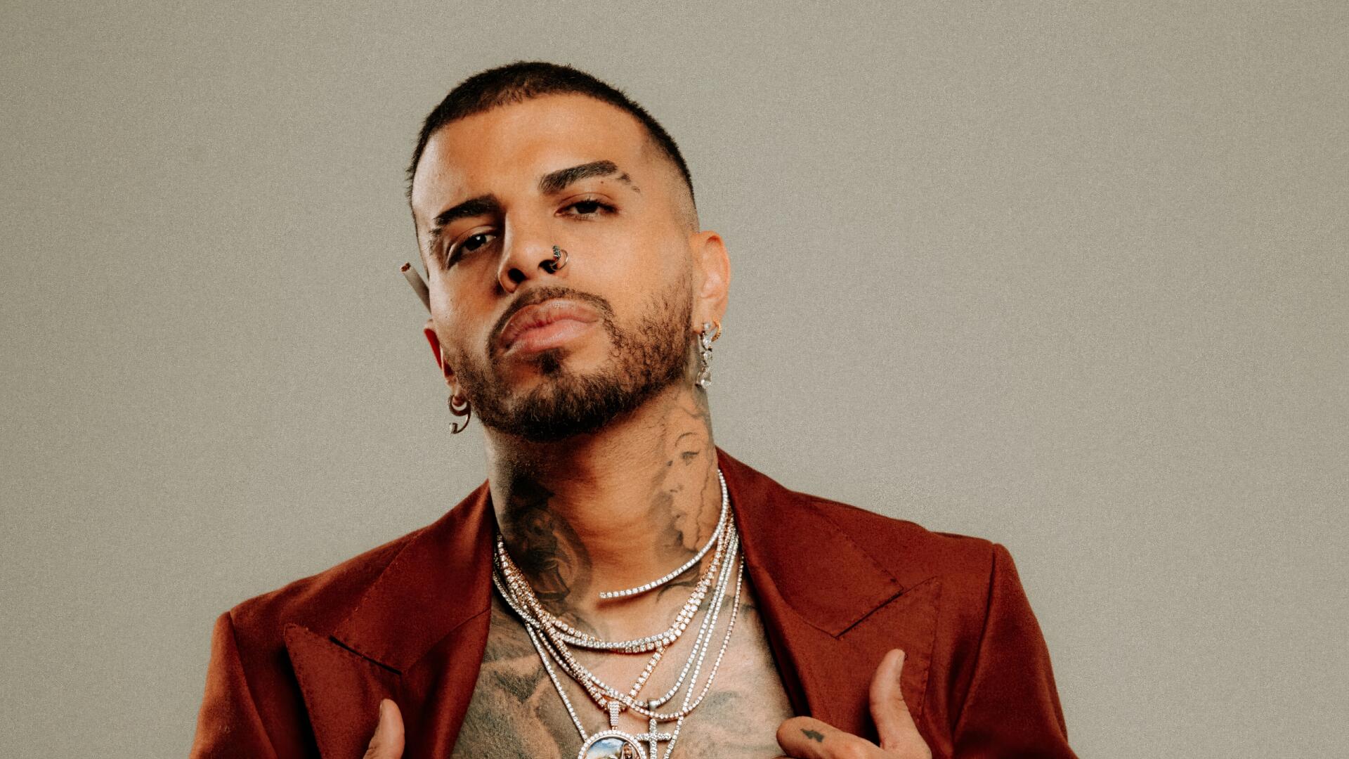HD PC desktop wallpaper featuring Rauw Alejandro, showcasing him in a confident pose with layered necklaces, earrings, and a maroon jacket, representing music and style.