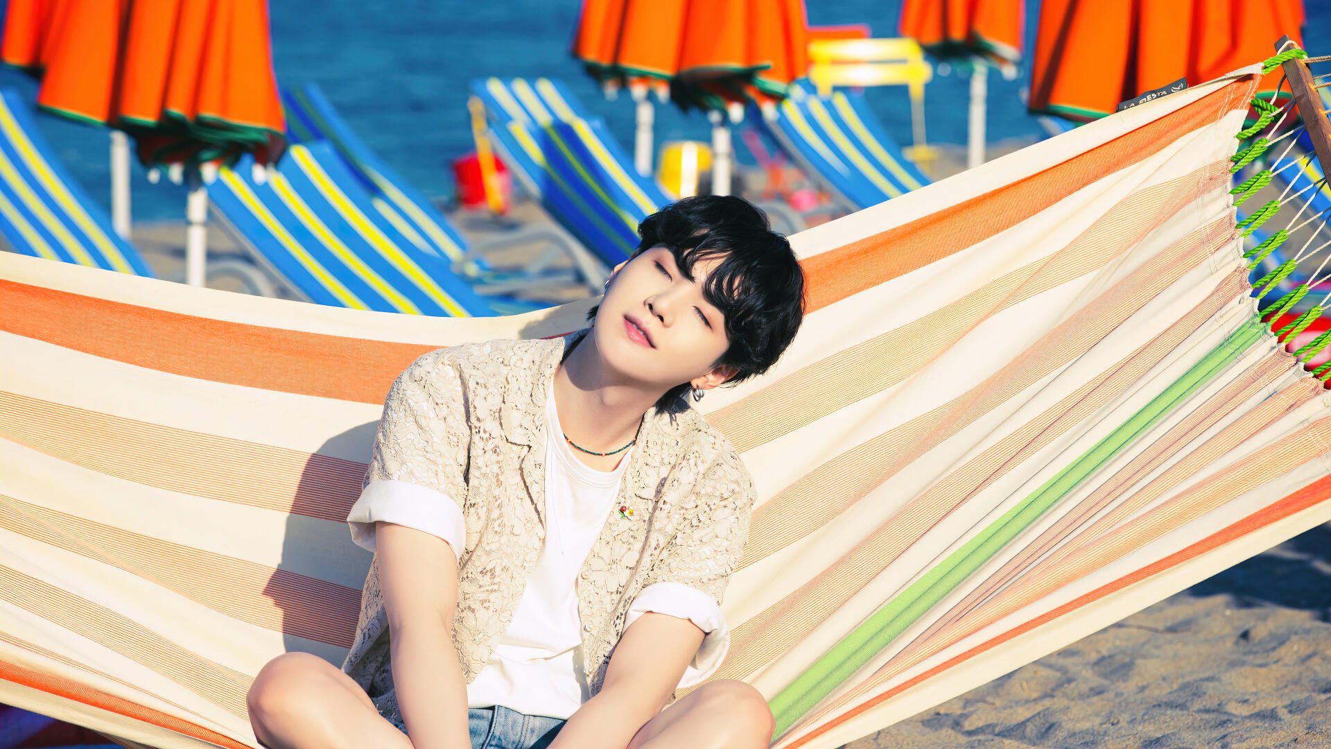 4K Ultra HD desktop wallpaper featuring BTS member Suga (Min Yoongi) relaxing on a striped hammock at a vibrant beach setting with colorful umbrellas in the background.