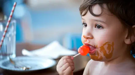 A young child enjoys a bright red lollipop, with playful expression and a smeared candy face, set against a softly blurred background, captured in vibrant 4K Ultra HD.